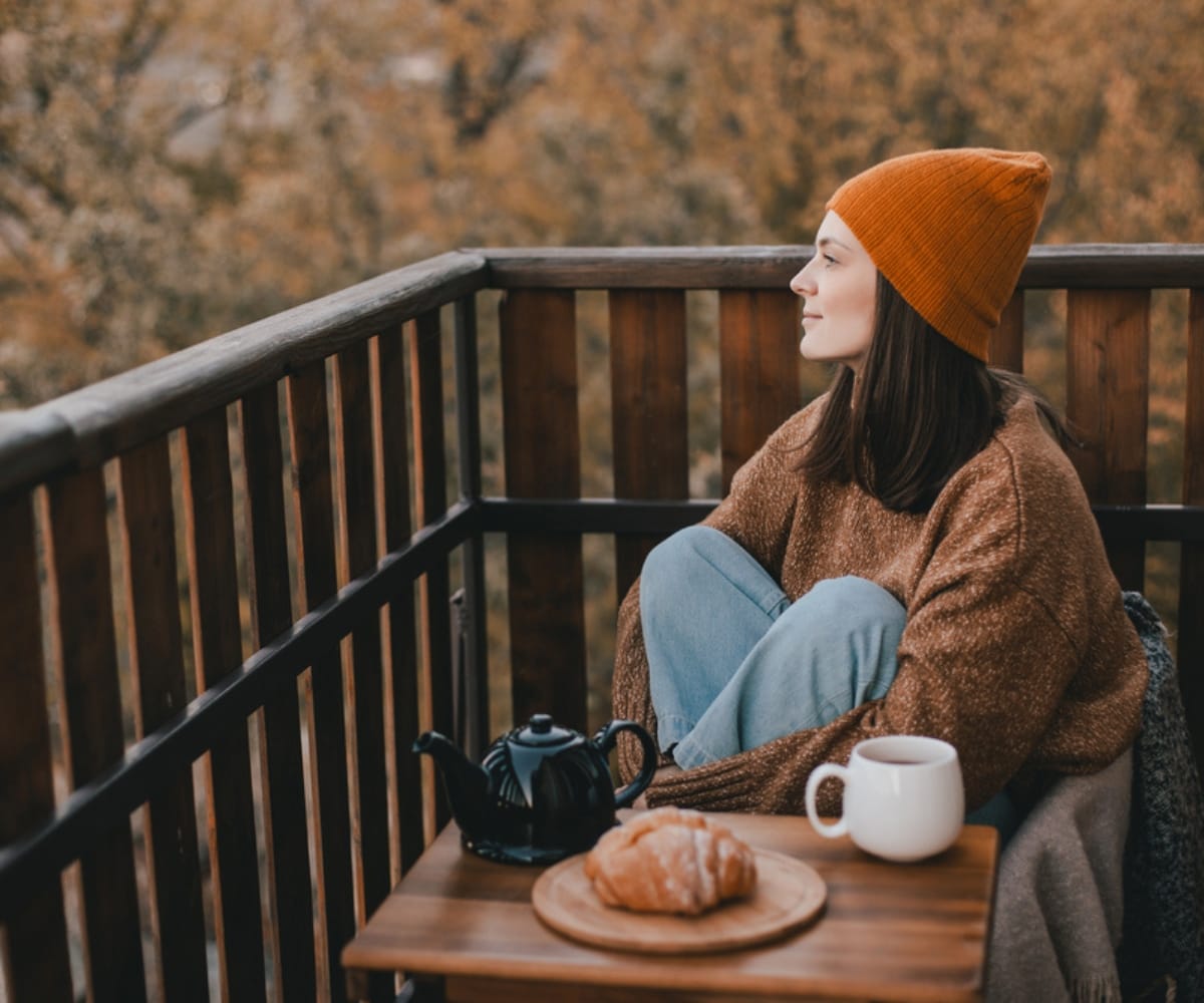 A young woman wearing a beanie and cardigan enjoys a croissant and coffee outside