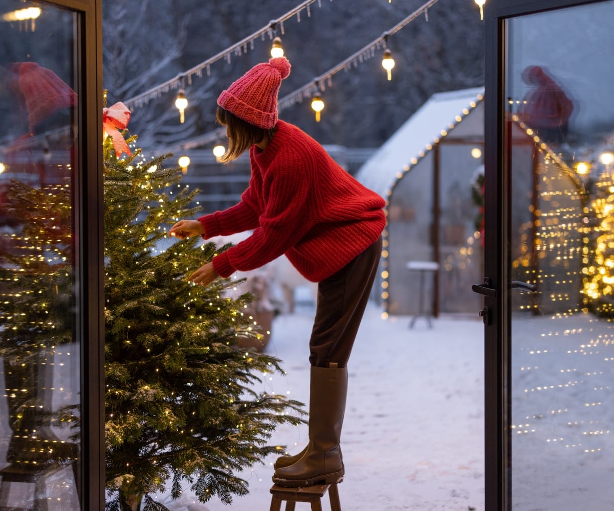 A woman decorates an outdoor christmas tree under an awning