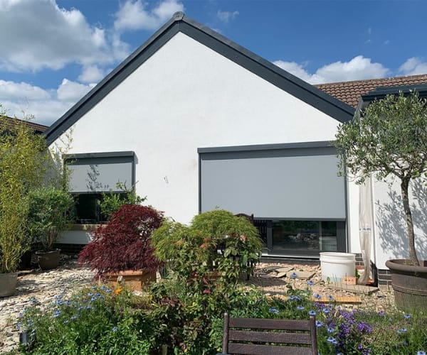 Rear garden of a house featuring two grey automated blinds from The Great British Awning and Blinds Company, with colourful plants in the foreground