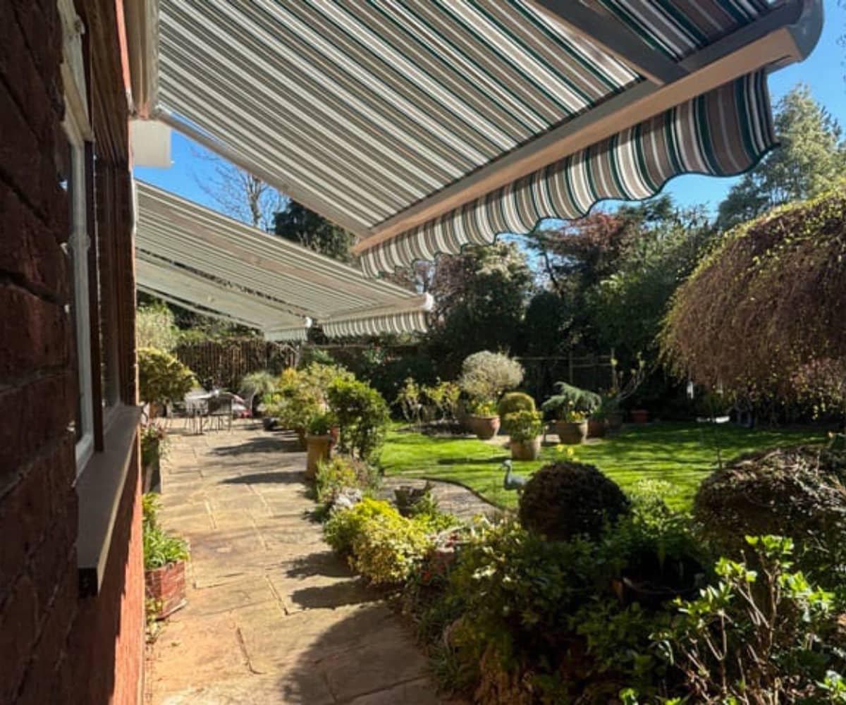 Striped garden awnings extended over a patio beside a red-brick house, shading a landscaped garden