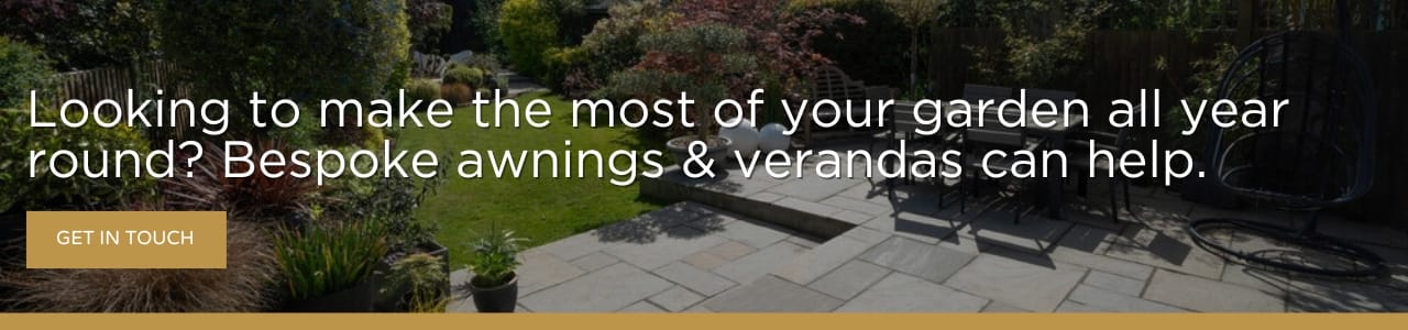 A landscaped garden with a paved patio area surrounded by lush greenery and plants. Outdoor furniture, including a table and chairs, sits in the sunlight beneath a message banner promoting garden awnings and verandas.