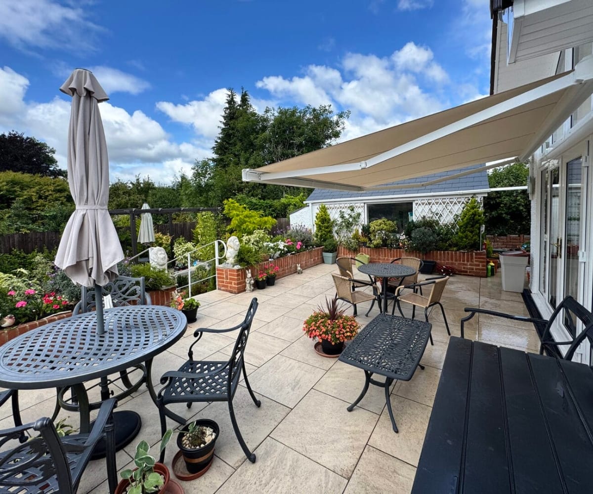 A bright patio area with multiple seating options and a large beige retractable awning extending from the house. The space is surrounded by potted plants, flowers, and brick planters, creating a peaceful, sunlit garden atmosphere.