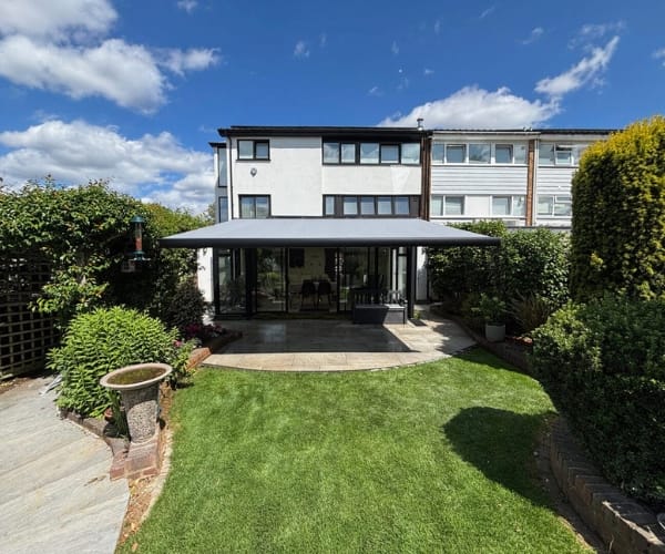 A modern home with a grey retractable awning extending over a stone patio area. The patio leads onto a well-kept garden with bright green grass and neat hedges on either side. The scene is set under a clear blue sky with scattered clouds.