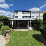 A modern home with a grey retractable awning extending over a stone patio area. The patio leads onto a well-kept garden with bright green grass and neat hedges on either side. The scene is set under a clear blue sky with scattered clouds.