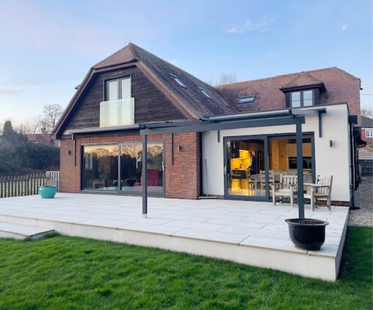 A modern detached home with a glass veranda extending over a tiled patio. The house features large sliding doors and wooden cladding under a pitched roof, with outdoor seating arranged neatly on the patio.