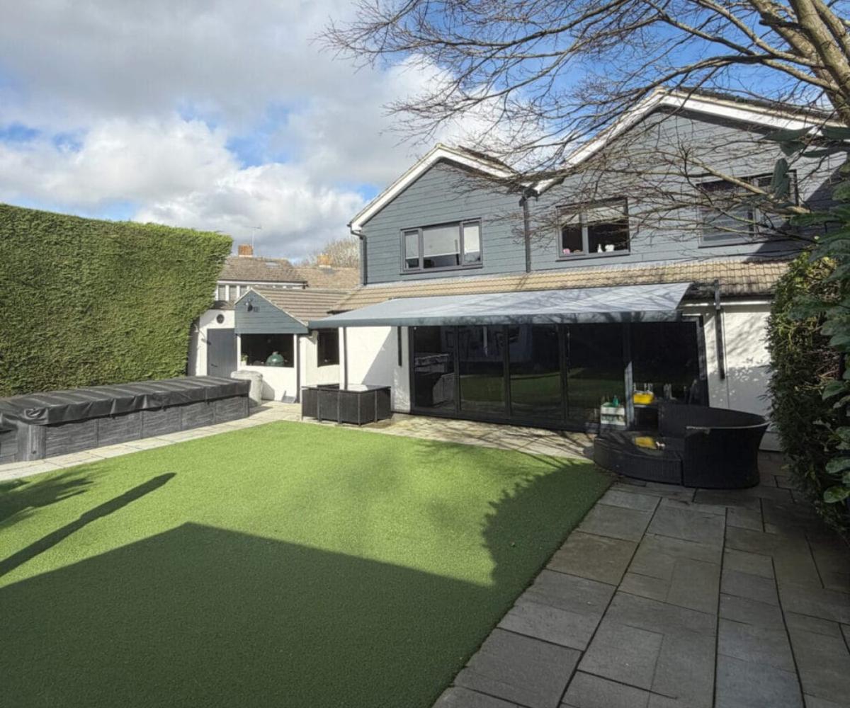 Awning quality: The rear of a grey rendered detached house with a large charcoal retractable awning extended over bi-fold doors, overlooking an artificial lawn, covered hot tub, and paved patio with rattan garden furniture.