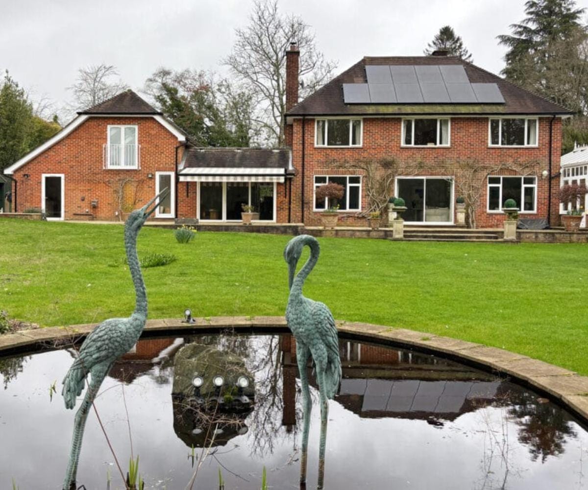 Awning comparison: The rear of a large red-brick detached house viewed across a formal garden pond, with two bronze heron sculptures at the water's edge, a well-maintained lawn, and a striped retractable awning extended over the rear extension.