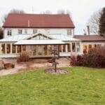 Luxury awnings: The same white-rendered detached house photographed from further back across a wide lawn, with the cream conservatory warmly lit from within, a white awning partially extended, and bare winter trees and shrubs surrounding the garden.
