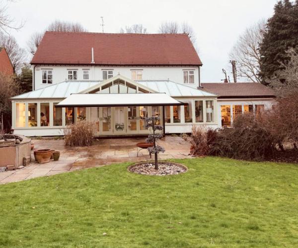 Luxury awnings (inline): The rear of a white-rendered detached house with a large cream-framed conservatory extension, a retracted white awning above the doors, a stone-paved patio with terracotta planters, and a sculpted topiary tree in the centre of the lawn, photographed in winter.