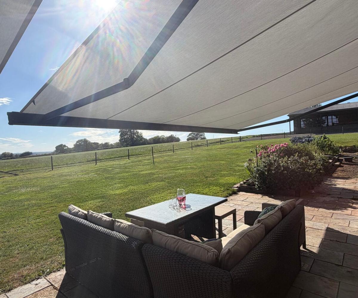 Outdoor dining at home UK A view from beneath a large cream retractable awning looking out over a wide rural landscape, with a grey rattan dining set on a sandstone patio in the foreground and open fields, fencing and flowering shrubs visible beyond.