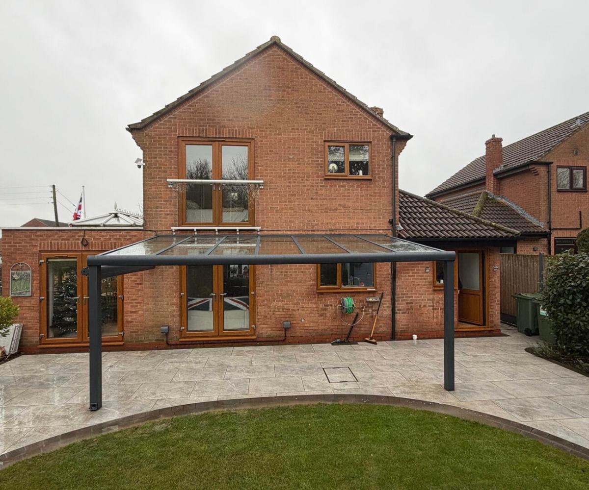 Shading a patio without losing the view The rear of a two-storey red brick house on an overcast day, with a freestanding glass-roofed pergola structure fitted against the back wall over a wide paved patio. Bi-fold doors open onto the patio, which is bordered by a curved lawn edge.