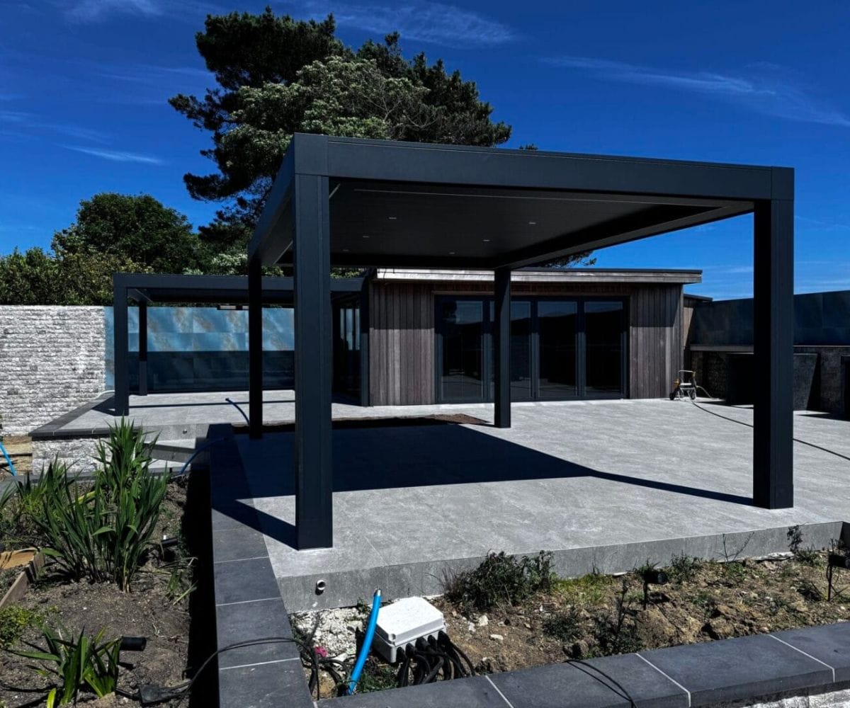 Extending your living space outdoors: A modern garden room with dark timber cladding and large glazed doors, set beneath a sleek black pergola on a grey stone-paved patio under a bright blue sky.
