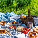 garden entertaining ideas for summer (featured) A sunny picnic spread laid out on a blue checked blanket on grass, featuring pretzels, bowls of fresh fruit and raspberries, cherry tomatoes, breadsticks, a jar of nuts, a round loaf of bread and a wicker basket filled with drinks.