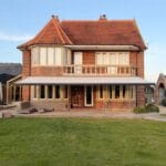Garden room ideas UK: The rear of a large traditional red-brick house with an extended canopy awning over the ground floor, a well-maintained lawn, and outdoor loungers on a paved terrace.