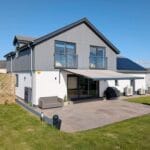 how to add shade to a garden without blocking light (featured) The rear exterior of a modern grey and white two-storey house on a sunny day, with a large retractable awning extended over a stone patio. A grey sofa, covered barbecue and air conditioning units are visible beside bi-fold doors leading inside.