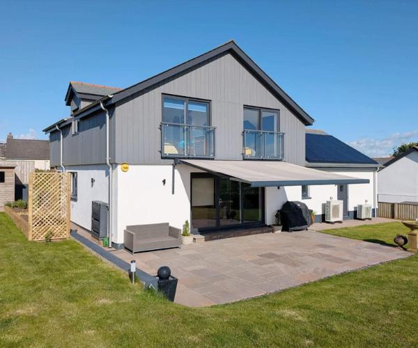 how to add shade to a garden without blocking light A rear view of a modern grey and white two-storey house under a clear blue sky, with a retractable awning extended over a stone-paved patio. A small grey sofa, a covered barbecue and air conditioning units sit alongside bi-fold doors, with a neatly kept lawn in the foreground.