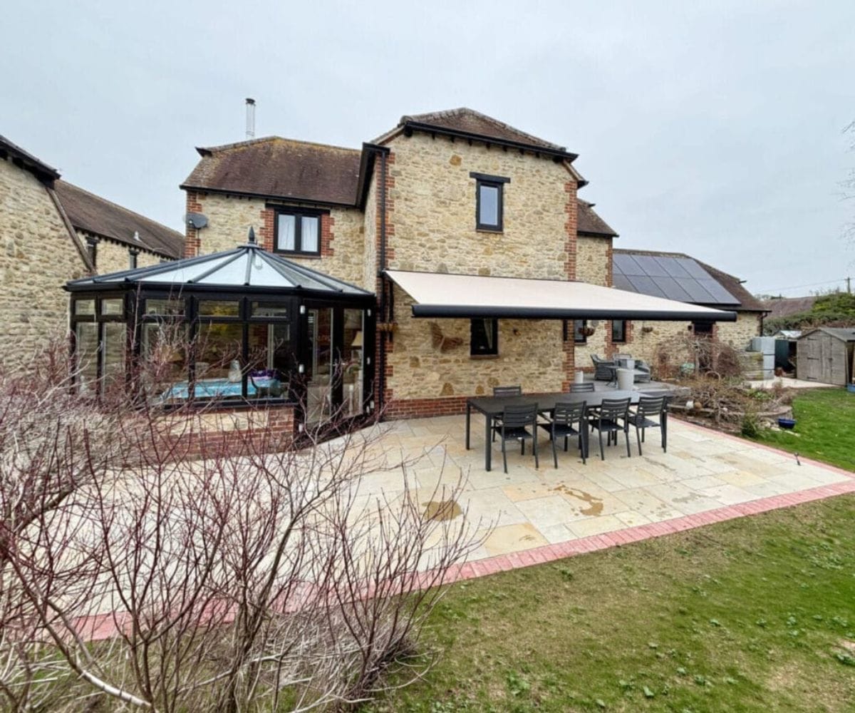 How to make your garden feel like an extra room: The rear of a stone and brick country house featuring a glass conservatory, a retractable cream awning over a paved patio, an outdoor dining set, and solar panels on the roof.