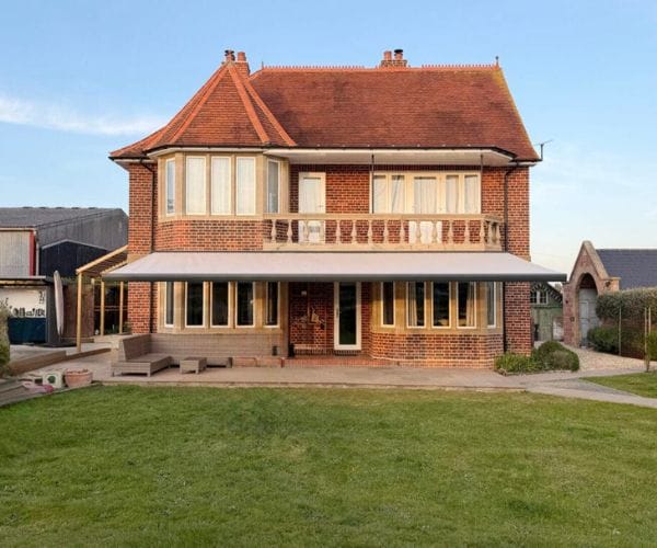 Outdoor rooms inline: The rear of a large traditional red-brick house with a retractable canopy awning extending over the ground floor, a well-maintained lawn, and outdoor loungers on a paved terrace.