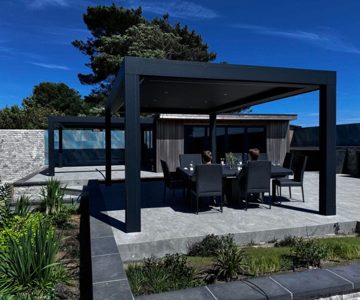 extending your living space outdoors: A black aluminium pergola over a grey stone patio with two people seated at a black dining table underneath, a wooden garden building and a stone wall in the background under a bright blue sky.