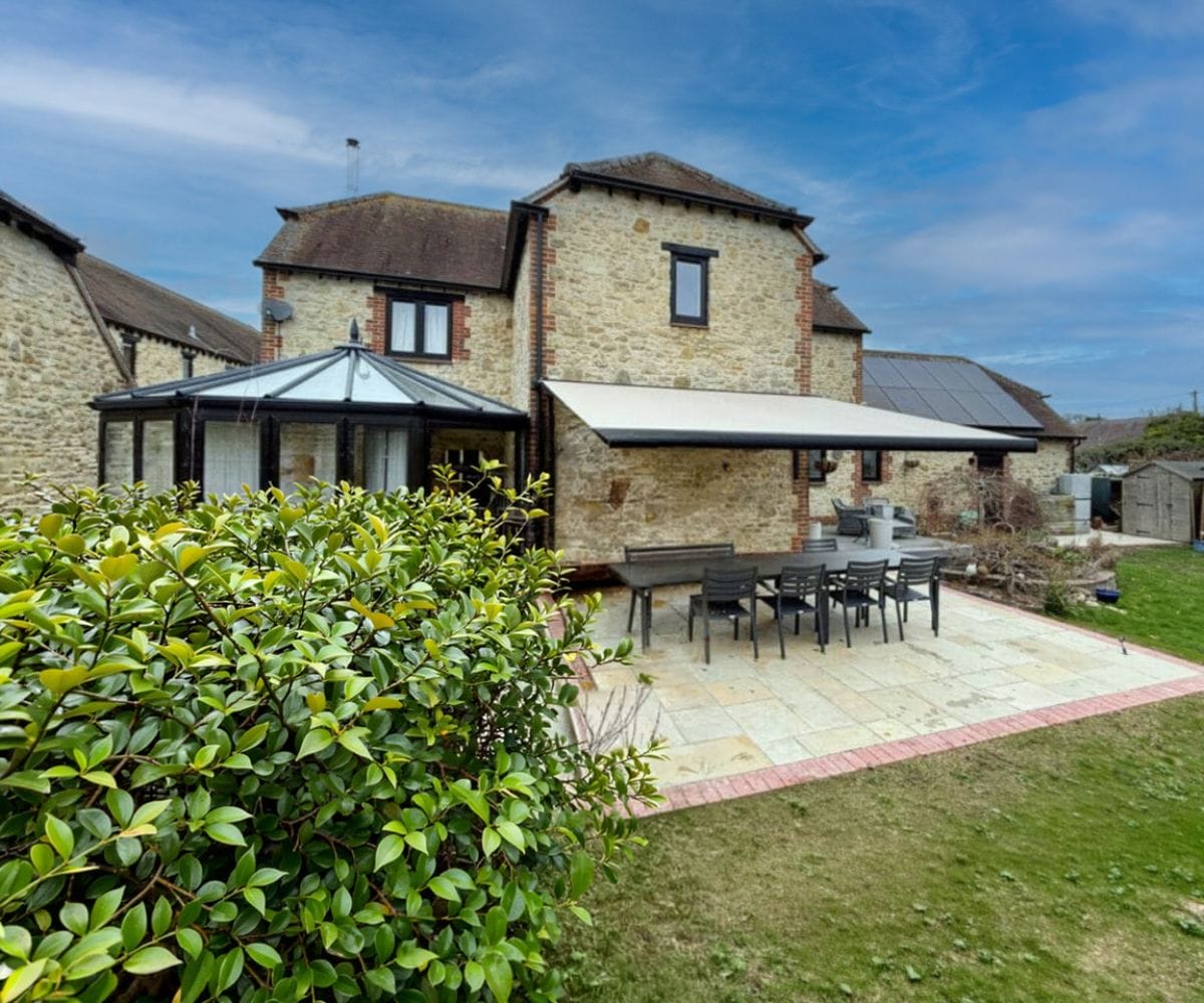 how to make your garden feel like an extra room: A stone cottage with a large white retractable awning extended over a black outdoor dining table and chairs on a paved patio, with a conservatory, solar panels and a landscaped garden surrounding the house.