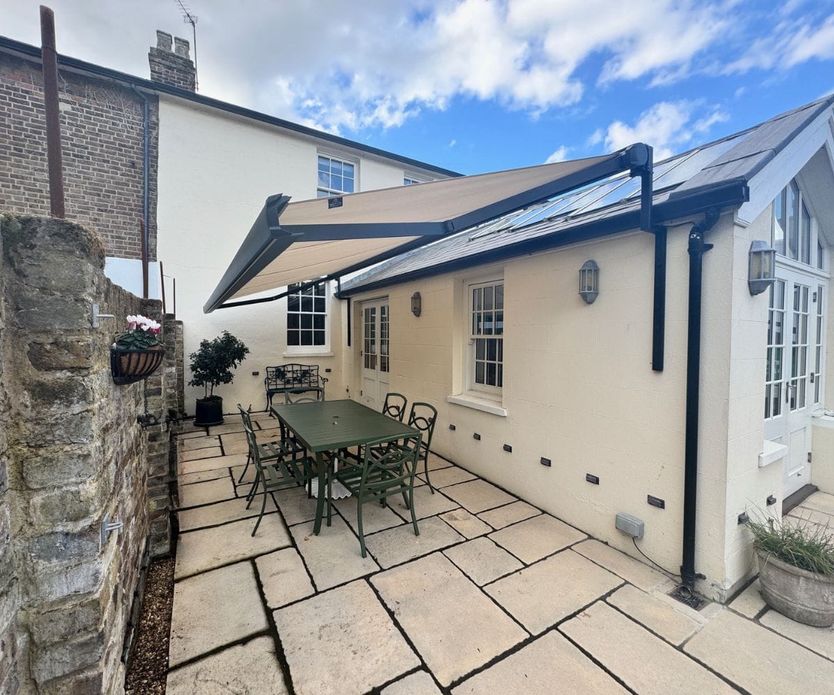 awning fixing brackets: A retractable fabric awning mounted on the exterior wall of a cream-rendered house, extending over a paved patio with a green metal table and chairs, beside a stone garden wall with a hanging flower basket.
