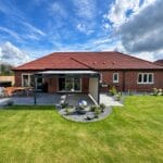 awning for bungalow: The rear of a red-brick bungalow with a grey retractable awning extending over a paved patio, set in a large lawned garden with a water feature, garden furniture and a hot tub under a blue sky.