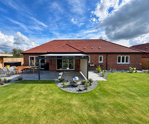awning for low wall: The rear of a red-brick bungalow with a grey retractable awning extending over a paved patio, set in a large lawned garden with a spherical water feature, garden furniture and a hot tub under a blue sky.
