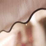 Can You Use an Awning in the Rain: Close-up of rain droplets running down a beige scalloped awning fabric, with a single drop falling from its dark trimmed edge.