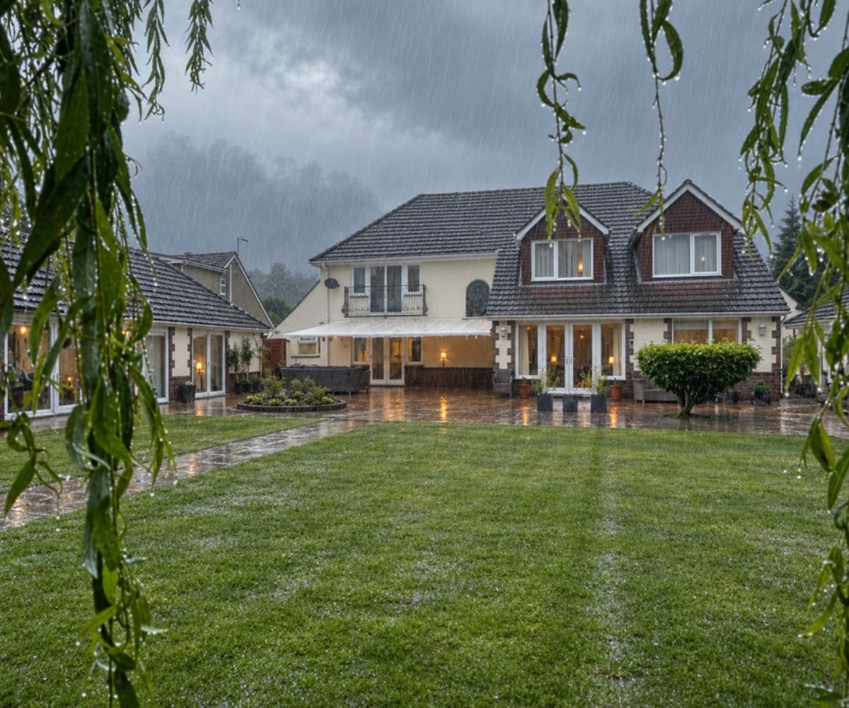A cream rendered house with a dark grey roof and a large patio awning extended over the rear terrace, viewed through hanging willow tree branches across a neatly mown lawn on a rainy, stormy winter day.