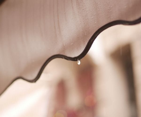 awning rain (inline): Close-up of rain droplets running down a beige scalloped awning fabric, with a single drop falling from its dark trimmed edge.